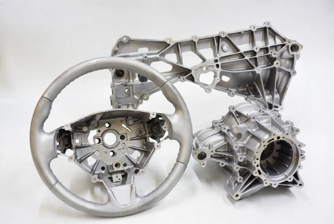 Die cast and CNC milled magnesium alloy ultra-lightweight automotive parts including steering wheels, seat frames, gearbox housings and instrument panels with silver-gray finish on clean white background
